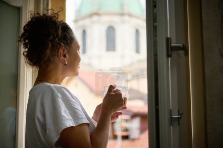Blick von hinten auf eine Touristin, die eine Tasse Aromakaffee in der Hand hält, ihren morgendlichen Espresso trinkt und aus dem Fenster ihres Hotelzimmers die Kathedrale bewundert. Menschen. Tourismus. Reisekonzept