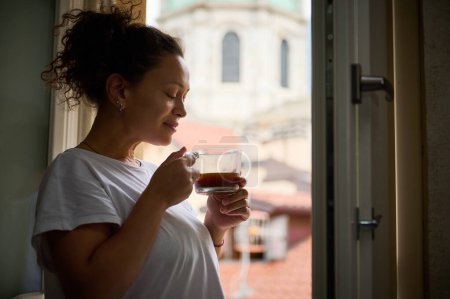 Glückliche multiethnische Frau mit geschlossenen Augen und einer Tasse frisch gebrühtem Kaffee am Fenster mit Blick auf die mittelalterliche Kathedrale Santa Maria di Assunta in der italienischen Stadt Como