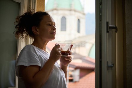 Entzückende junge multiethnische Frau genießt ihren Morgenkaffee und steht am Fenster mit Blick auf die mittelalterliche Kathedrale Santa Maria di Assunta in der italienischen Stadt Como.