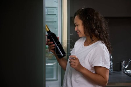 Photo for Side portrait of a dipsomaniac, alcoholic drunk young woman standing at the refrigerator in the home kitchen, choosing a bottle of wine in the morning. Alcoholism, mental health and social problems - Royalty Free Image