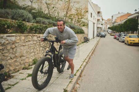 Foto de Hombre joven caucásico de 40 años, empujando bicicleta de montaña eléctrica por la calle. Estilo de vida activo. Deporte. Viaje de aventura en bicicleta. Utilizar el modo de transporte urbano ecológico de la bicicleta - Imagen libre de derechos