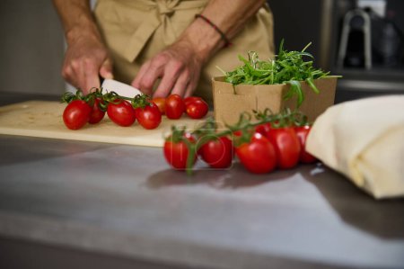 Téléchargez les photos : Vue rapprochée des mains d'un chef masculin hachant une cerise de tomate sur une planche à découper. Ingrédients sur la table de cuisine. Homme cuisine salade saine dans l'intérieur de la cuisine maison minimaliste. Concept alimentaire - en image libre de droit