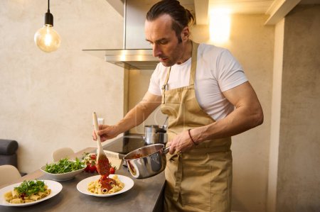 European professional chef plating up pasta before serving it to the customer. Handsome man in apron, holding stainless steel saucepan and pouring tomato sauce on pasta, cooking dinner at home kitchen