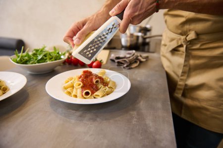 Close-up view of chef plating up pasta before serving it to the customer. Male hands holding grater, grating parmesan cheese, seasoning Italian pasta spaghetti while cooking dinner at home kitchen