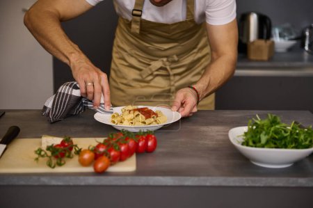 Close-up chef in beige apron, using kitchen towel, removing traces of tomato sauce on the white plate with Italian pasta before serving the dish. Fresh organic ingredients on the kitchen countertop