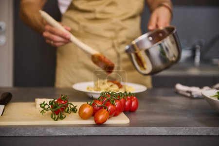 Focus on a branch of fresh ripe organic tomato cherry on the cutting board against the backdrop of blurred male chef cooking Italian pasta at home kitchen. Traditional Italian cuisine. Food background