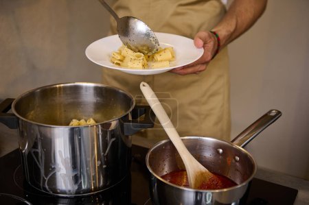 Close-up chef's hands plating up freshly cooked pasta, putting boiled penne on a white plate, standing at induction electric stove with saucepan full of tomato pasta. Italian culinary. Food background