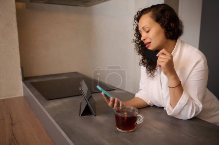 Belle jeune femme vérifiant les messages sur tablette numérique, assis sur une tasse de tisane chaude. Brune aux cheveux bouclés multi ethnique en peignoir blanc lisant les nouvelles sur smartphone, naviguant sur le web