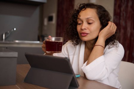 Attraktive junge brünette Frau liest Nachrichten auf einem digitalen Tablet, surft im Internet, sitzt bei einer Tasse Tee am Küchentisch und trägt einen weißen Bademantel