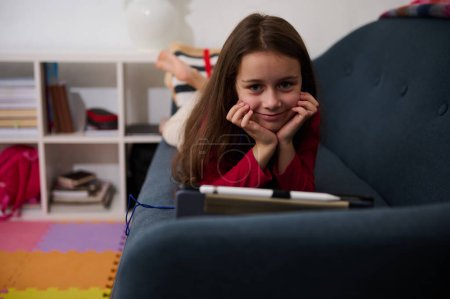 Little child girl, addicted to digital gadgets, playing online video games while lying on the sofa at home. Lovely school girl watching movies, smiling looking at camera. Happy carefree childhood