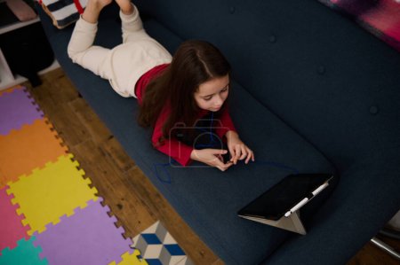 View from above of a Caucasian cute school girl lying on a sofa and using digital tablet at home. Kids education and entertainment. Childhood, Digital gadget addiction problems