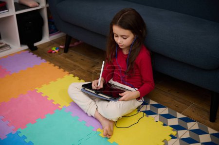 Kid girl doing homework on tablet at home, sitting on multicolored puzzle carpet. People. Online communication. Distance learning and kids education concept. Copy ad space on black mockup touch screen