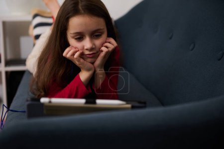 Großaufnahme kaukasisches Grundschulmädchen, ein entzückendes kleines Mädchen, das an ihrem smarten digitalen Tablet Spaß hat, während es an ihrem Wochenende auf der Couch liegt. Kleines Kind schaut Zeichentrickfilm auf digitalem Gerät