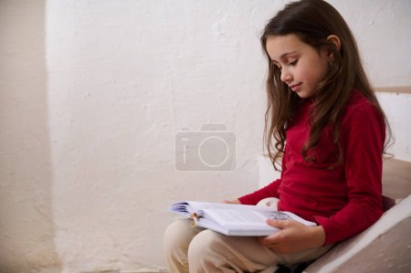 Photo for Authentic portrait of an adorable little kid girl in white sports trousers and red shirts, reading a book, sitting on the steps at home, over white wall background. Copy space for advertisement - Royalty Free Image