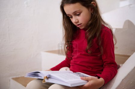 Authentic portrait of smart little kid girl leafing through the pages of a hardcover book, reading a book, sitting on the steps at home, over white wall background of a rural house. Copy space for ads