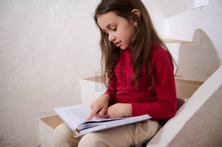 Photo for Caucasian cute child, elementary age smart little kid girl leafing through the pages of a hardcover book, reading a fairytale, sitting on the steps at home, over white wall background of a rural house - Royalty Free Image