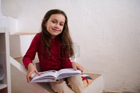 Photo for Smart elementary school student girl smiles at camera, sitting on steps at home with a book in hands. Cute child reading book. World Book Day concept. Back to school on new semester of academic year - Royalty Free Image