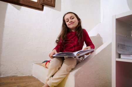 Entzückendes, smartes Grundschülermädchen, das lächelnd in die Kamera blickt, zu Hause auf den Stufen sitzt und ein Buch in der Hand hält. Intelligentes kleines Mädchen beim Lesen eines Buches. Welttag des Buches. Zurück zur Schule