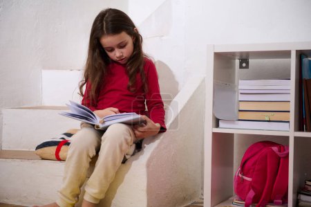 Kaukasisches Mädchen im Schulalter liest in gemütlicher häuslicher Atmosphäre ein Buch und sitzt auf den Stufen ihres Zimmers. Das Konzept der Rückkehr zur Schule. Intelligente Kinder, Erudition und Lernen. Welttag des Buches