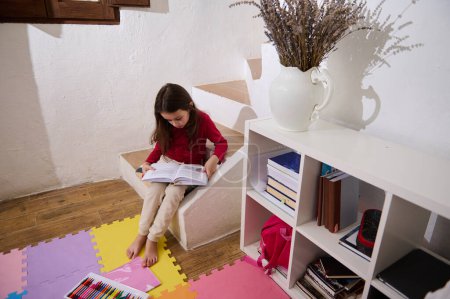 Photo for View from above on erudite child girl, elementary age schoolgirl reading a book in her room. Nearby bookshelf with books and school supplies. multi colored puzzle carpet on the floor. Autism concept - Royalty Free Image