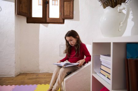 Photo for Authentic portrait of erudite child girl, elementary age schoolgirl reading book in her room over window background. Nearby bookshelf with books and school supplies. Smart kids. World book Day - Royalty Free Image