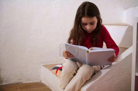 Photo for Caucasian cute little girl sitting on steps at home, reading a book over white wall background. Literature. Erudition. Knowledge. World Book Day. Back to school concept. Copy advertising space - Royalty Free Image