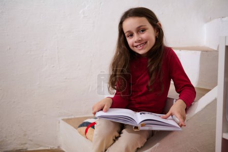 Photo for Authentic portrait of an adorable child girl holding a book, smiling looking at camera, sitting on steps at home over white wall background. Kids education and entertainment. Back to school - Royalty Free Image