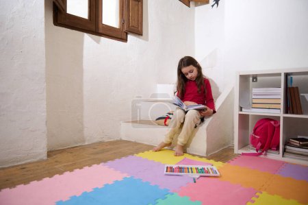 Photo for Smart elementary age girl leafing through the pages of a book, reading a fairytale, sitting on the steps barefoot on multicolored puzzle carpet at home, over white wall background of a rural house. - Royalty Free Image