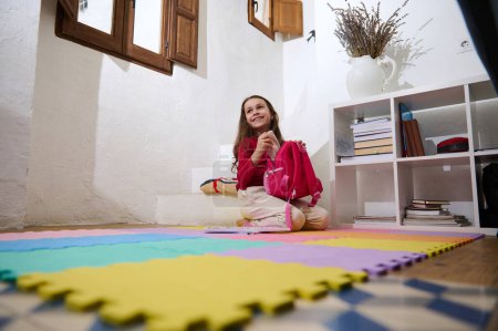 Kaukasische kleine Mädchen packen ihren Rucksack, sitzen auf bunten Puzzle-Teppich zu Hause, lächeln und schauen weg. Menschen. Lebensstil. Kinder. Bildung. Zum neuen Semester zurück in die Schule