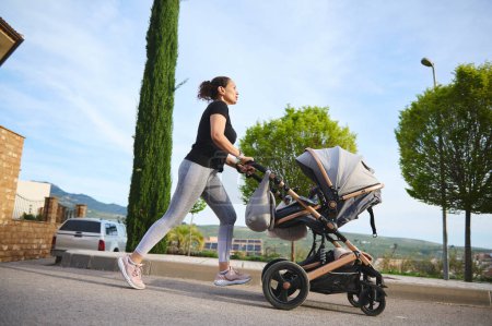 Ganztägige Aufnahme einer glücklichen Frau, die morgens joggt, einen Kinderwagen auf der Straße schiebt und ihren aktiven Lebensstil während des Mutterschaftsurlaubs genießt. Menschen. Mutterschafts- und Kindheitskonzept