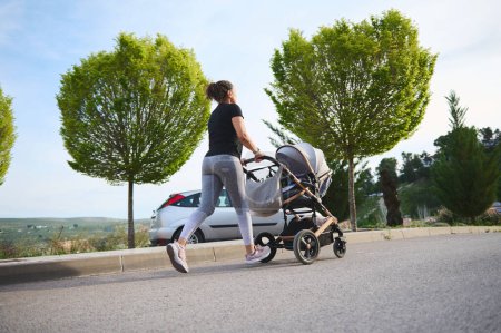 Blick von hinten auf eine junge Frau, die mit dem Kinderwagen ihres Babys im Freien joggt, um nach der Geburt fit zu bleiben.