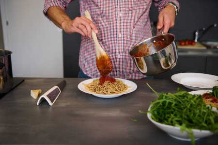Cropped image of chef pouring tomato sauce on Italian pasta, plating up the dish before serving. Man cooking spaghetti for family dinner at home kitchen. Cuisine. Culinary. Epicure. Food concept