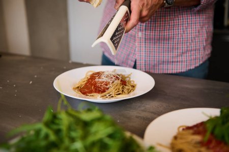 Männliche Hände in Großaufnahme reiben Käse auf frisch gekochten italienischen Spaghetti mit Tomatensauce und bereiten das Familienessen in der heimischen Küche zu. Italienische Küche. Frische Zutaten auf dem Küchentisch