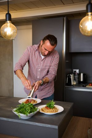 Vertical shot of young adult man grating cheese, seasoning the plate with freshly cooked meal, according to traditional Italian recipe, standing at kitchen counter in minimalist home kitchen interior