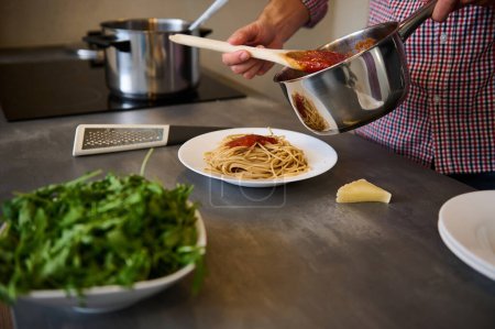 Young man cooking Italian pasta for dinner in the home kitchen. Close-up male hands holding a steel saucepan, pouring tomato passata sauce freshly cooked spaghetti,. seasoning the dish before serving