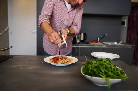 European man cooking pasta for dinner at home. Close-up hands holding a grater, grating cheese while seasoning the freshly boiled spaghetti, preparing healthy meal according to Italian recipe