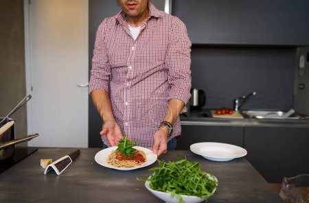 Close-up view of a young man cooking Italian pasta spaghetti for dinner in the home kitchen. Focus on served plate with Italian spaghetti with tomato sauce and greens arugula herbs