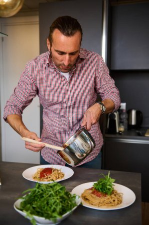 Attractive handsome European man pouring tomato sauce on spaghetti pasta, plating up the dish before serving. Man cooking dinner at home kitchen. Italian Cuisine. Culinary. Epicure. Food concept