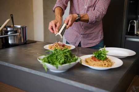 Close-up man cooking Italian spaghetti for dinner in stylish modern minimalist home kitchen interior, grating parmesan cheese over a plate with freshly boiled pasta with tomato sauce