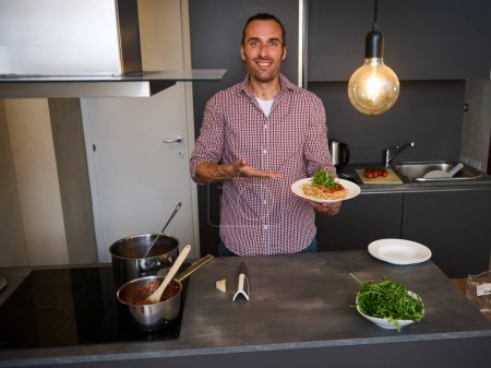 Attractive smiling young man 40s, showing at camera a plate with freshly cooked spaghetti pasta, standing at kitchen table with fresh ingredients and kitchen utensils. People. Italian food and culture