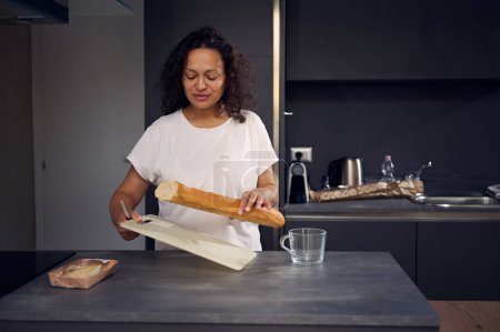 Authentisches Porträt einer jungen Frau mit einem Laib Brot und Schneidebrett, die zu Hause am Küchentisch steht und morgens mit der Zubereitung des Frühstücks beginnt. Das häusliche Leben. Menschen und Lebensstil