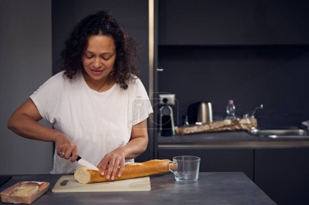 Hübsche Frau im weißen T-Shirt, steht am Küchentisch, hält ein Küchenmesser in der Hand und schneidet am Schneidebrett Brot, bereitet morgens in der heimischen Küche das Frühstück zu.