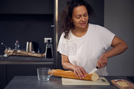 Gemischte Rasse schöne Frau in weißem T-Shirt, steht am Küchentisch, hält Messer in der Hand und schneidet einen Laib Vollkornbrot auf Schneidebrett, bereitet morgens das Frühstück in der heimischen Küche zu