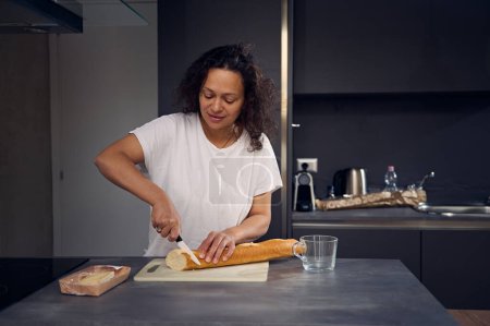Gemischte Rasse schöne Frau in weißem T-Shirt, steht an der Küchentheke, hält Messer und schneidet einen Laib französisches Baguette auf einem Schneidebrett, bereitet morgens das Frühstück in der heimischen Küche zu