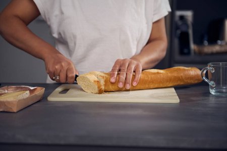 Frauenhände in Großaufnahme mit dem Küchenmesser, Brotlaibe und französisches Baguette auf Schneidebrett schneiden, Toastbrot für das Frühstück in der minimalistischen Wohnküche zubereiten
