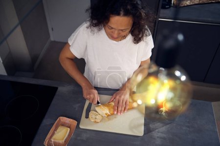 Draufsicht durch stylische Vintage-Lampe auf glückliche junge Frau, Hausfrau steht am Küchentisch und schneidet Brot auf Schneidebrett, macht Sandwiches mit Käse für den Imbiss zu Hause