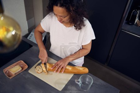 Blick von oben auf eine Frau, die ein Küchenmesser benutzt, einen Laib französisches Baguette-Vollkornbrot auf dem Schneidebrett schneidet, leckere Sandwiches mit Käse für das Frühstück in der heimischen Küche zubereitet.