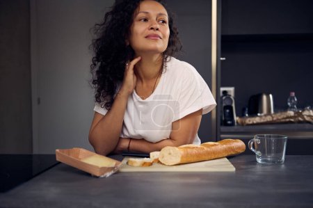 Authentisch schöne, lockige, brünette Frau in weißem T-Shirt, verträumt zur Seite schauend, mit einem Laib frischem Brot und einer Scheibe Käse zum Frühstück an der Arbeitsplatte in der Küche stehend. Menschen. Lebensstil