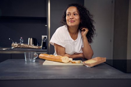 Lockige, multiethnische, brünette Frau in weißem T-Shirt, verträumt zur Seite blickend, mit einem Laib Brot und Scheiben Käse zum Frühstück am Küchentisch stehend. Menschen. Lebensstil