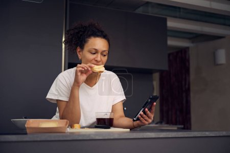 Belle femme multi ethnique 40s, manger un sandwich au fromage et vérifier le contenu des médias sociaux sur son smartphone, debout au comptoir de la cuisine dans l'intérieur minimaliste de la cuisine de la maison.